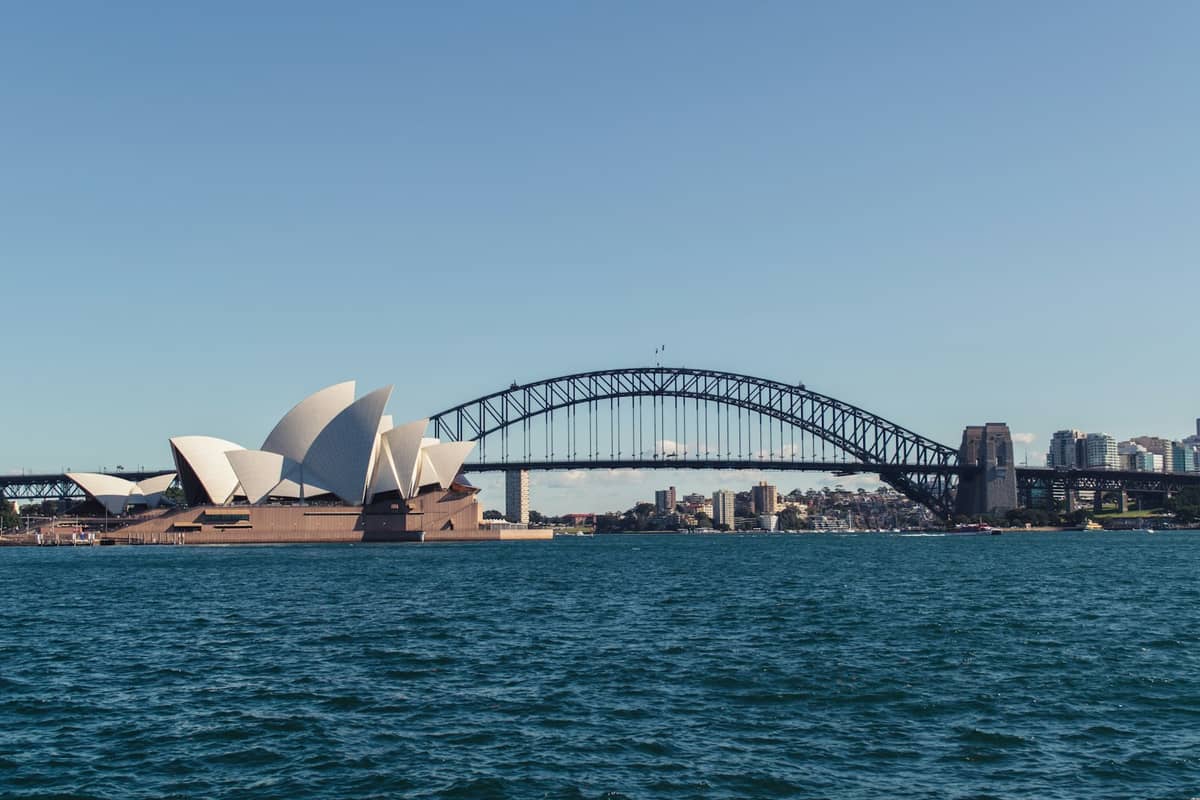 Opera w Sydney i Most Harbour Bridge obok siebie w słoneczny dzień – błękitne niebo i woda na pierwszym planie.