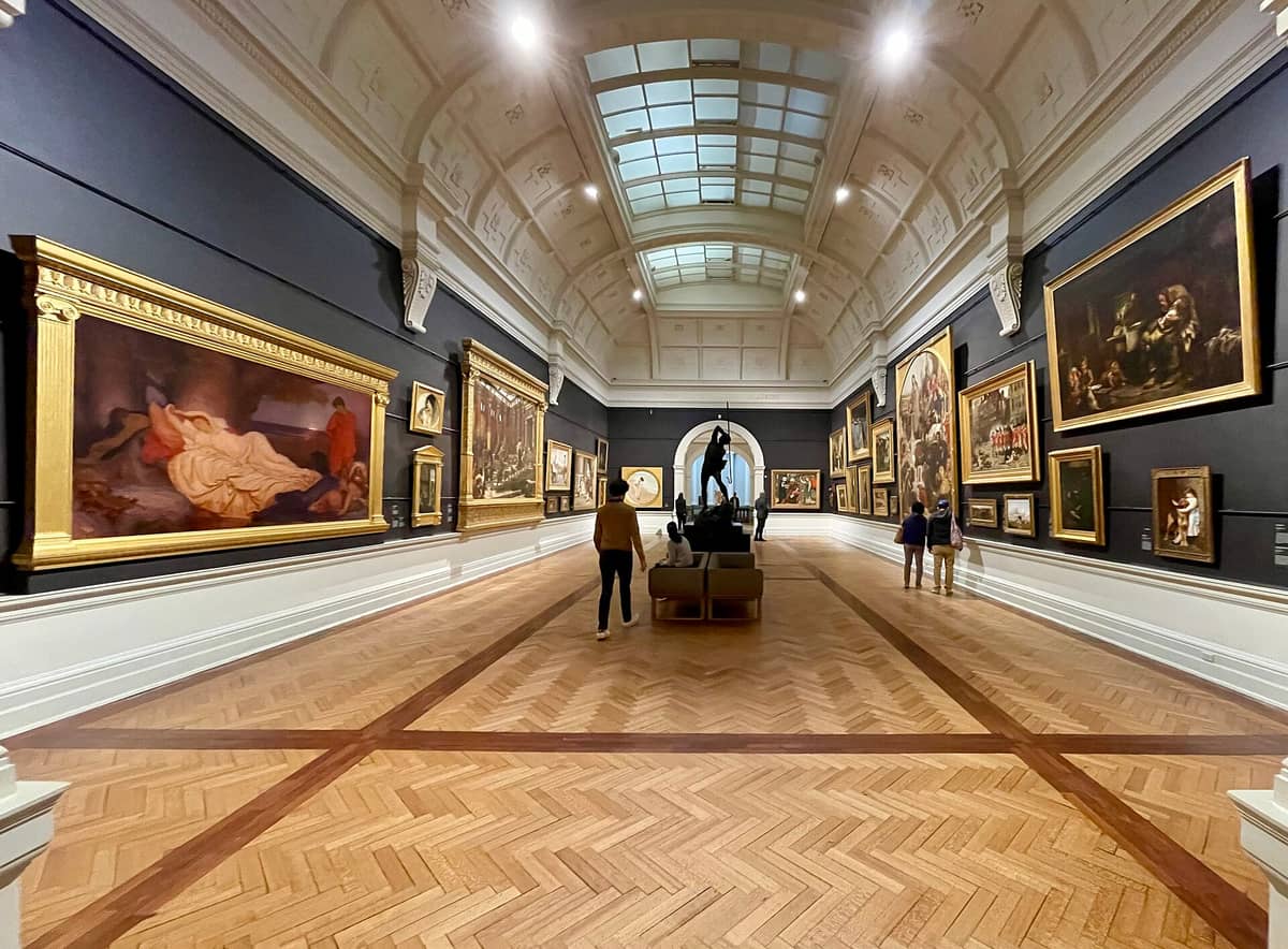 Wide view inside an art gallery with parquet flooring, ornate arched ceiling, and large classical paintings on display walls.