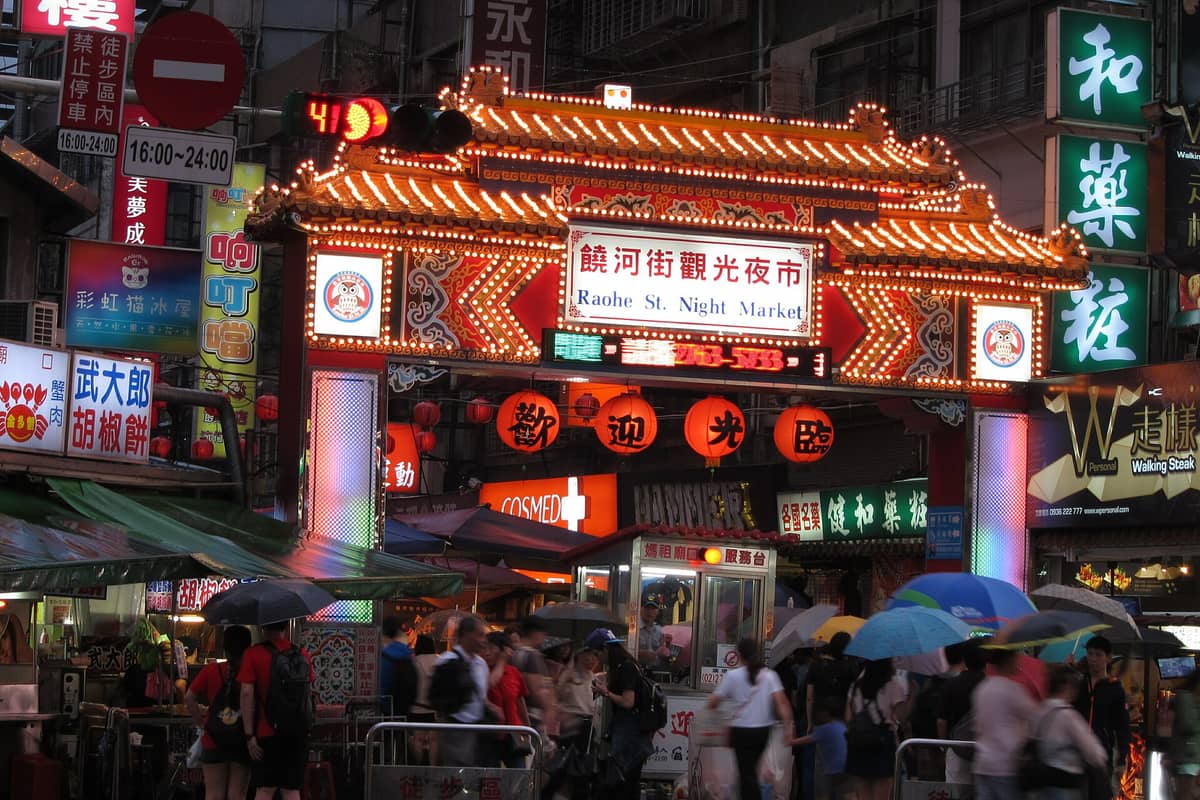 Überfüllter Eingang zum Raohe Street Nachtmarkt in Taipei, beleuchtet mit dekorativen Laternen, Neonlichtern und Marktschildern.