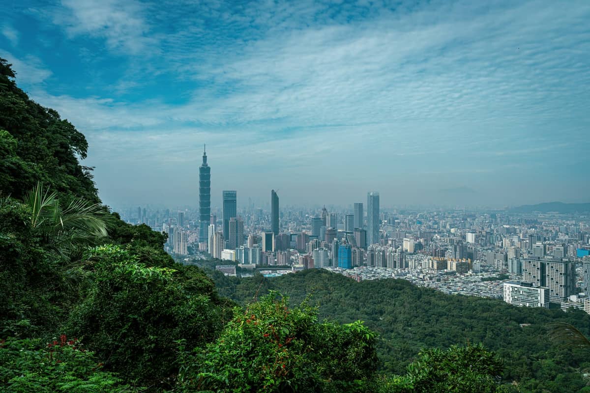 Blick von einem üppig grünen Hang über die Stadt Taipei und Taipei 101, unter einem blauen Himmel mit vereinzelten Wolken.