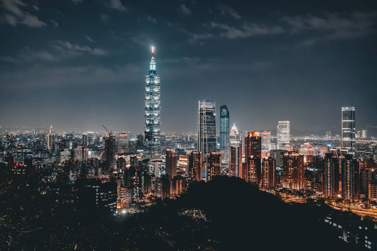 Nächtliches Panorama der Taipeh-Innenstadt mit beleuchtetem Taipei 101 und der Skyline der Stadt, aufgenommen von einem erhöhten Aussichtspunkt mit Grünflächen im Vordergrund.