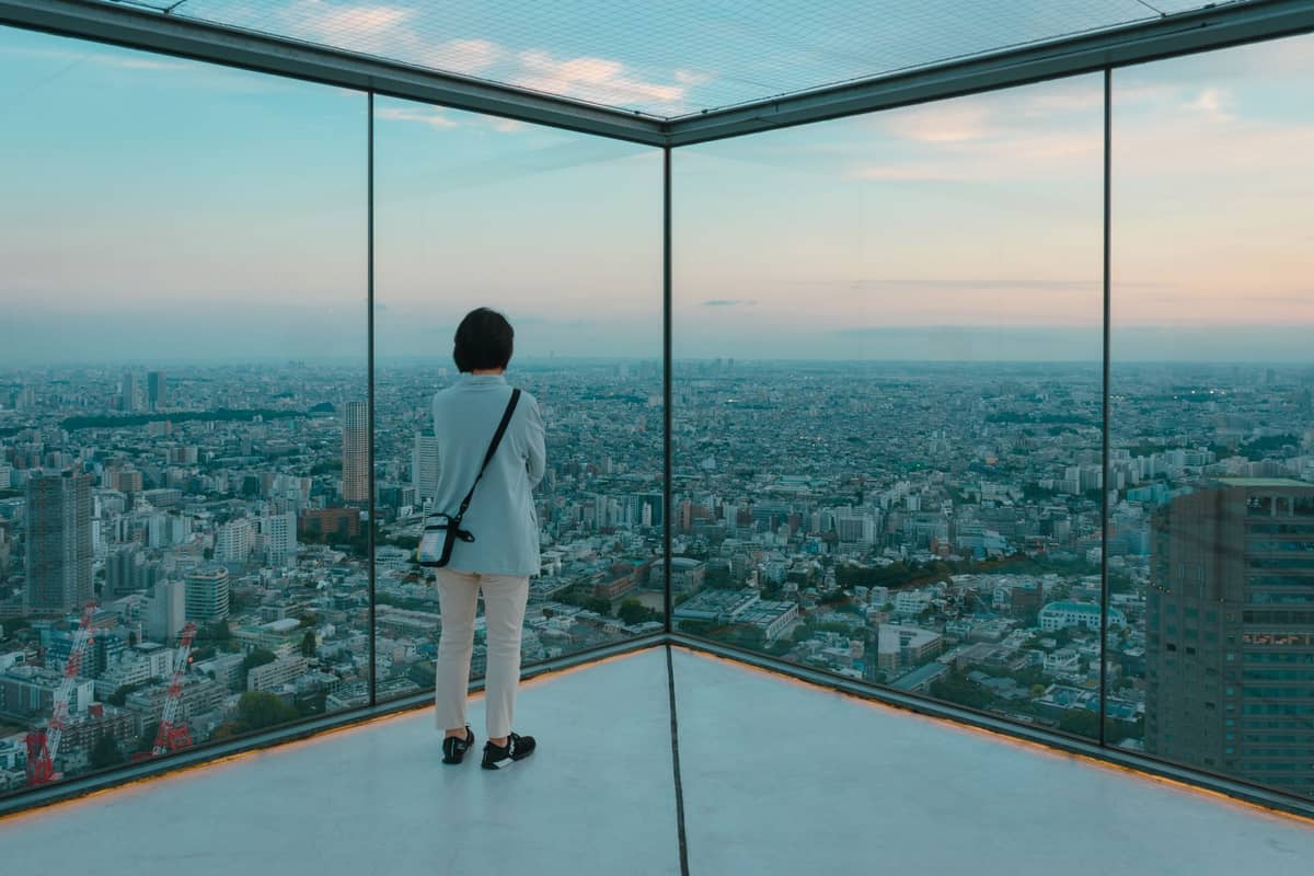 Woman stands alone at the corner of a modern glass observation deck, overlooking the Tokyo skyline in soft daylight.