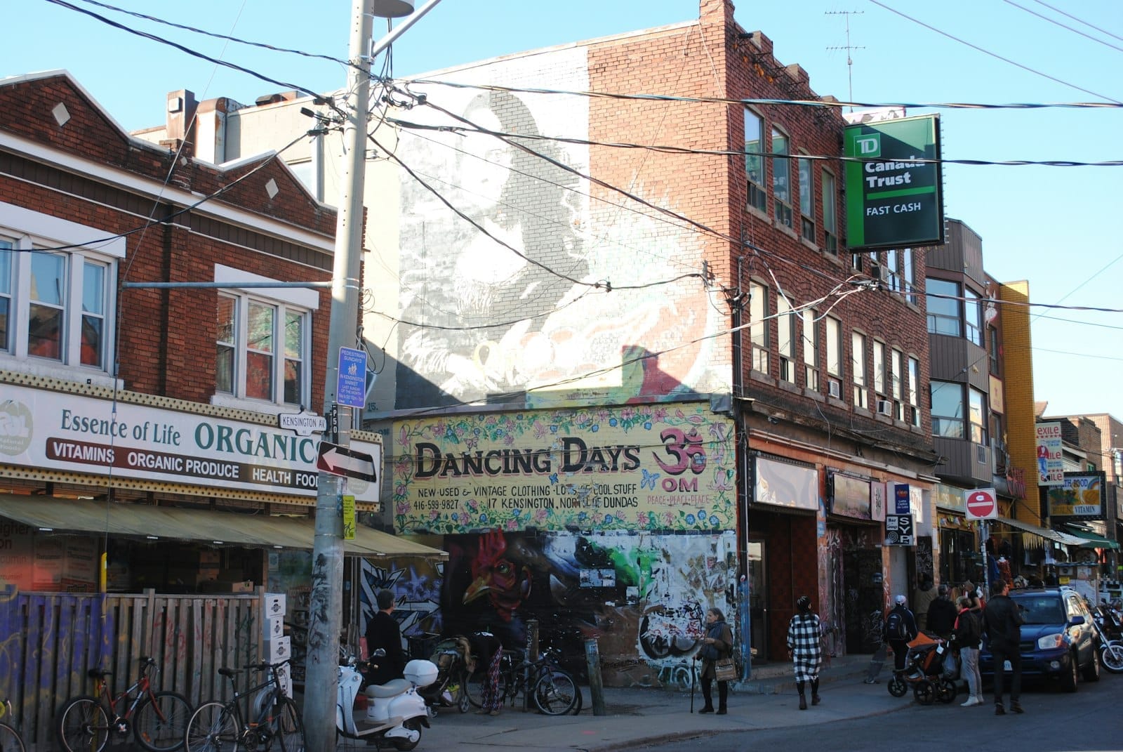 Vista da rua Kensington Market em Toronto, mostrando lojas independentes vibrantes, pedestres e um mural colorido em um dia ensolarado.