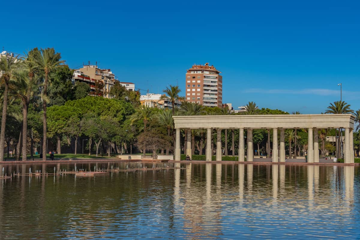 Grande stagno con una struttura a colonne e palme in un parco urbano, con edifici visibili sullo sfondo sotto un cielo azzurro limpido.