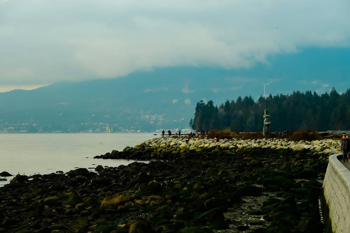 ทางเดินริม Seawall ที่โค้งไปตามแนวชายน้ำของ Vancouver พร้อมชายหาดหินกรวด รูปปั้น Inukshuk และภูเขาในฉากหลังใต้ท้องฟ้ามีเมฆ