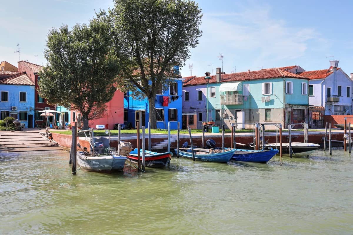 Bunte Häuser und kleine Boote entlang der Uferpromenade auf einer der Laguneninseln bei Venedig, mit Bäumen und ruhigem Wasser.