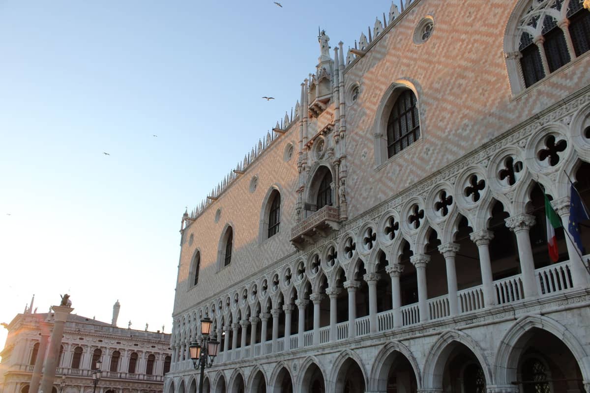 Weitwinkelansicht der verzierten gotischen Außenfassade des Dogenpalastes in Venedig, mit Bogenarkaden und dekorativen Fenstern bei Sonnenuntergang.