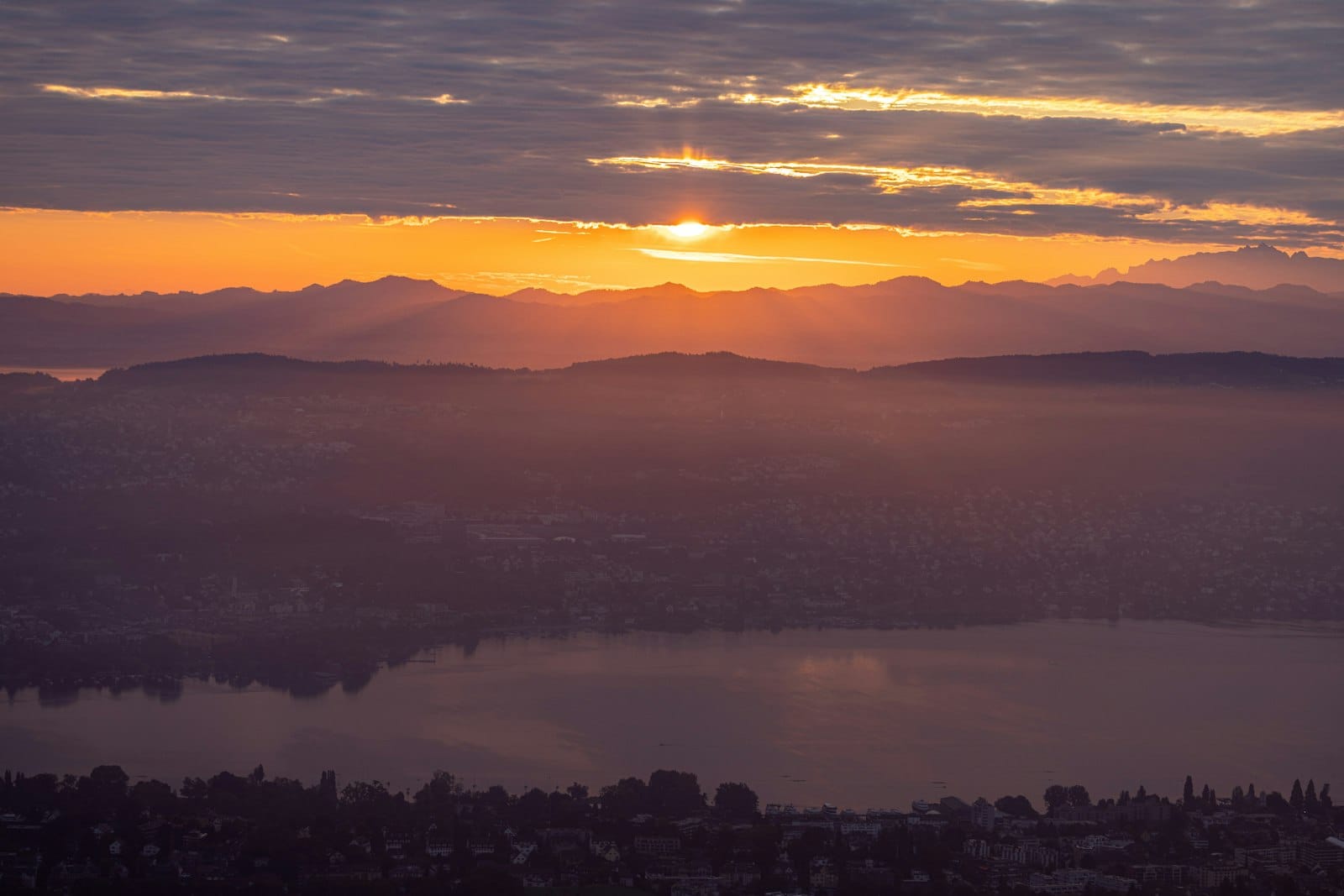 วิวกว้างจากมุมสูงเหนือเมือง Zurich ช่วงพระอาทิตย์ขึ้นหรือตก พร้อมทะเลสาบ Zurich ตัวเมือง และเทือกเขาไกลๆ ที่ถูกแสงทองส่องสว่าง