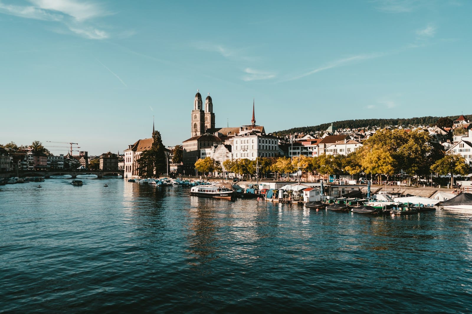 วิวเมืองซูริกริมแม่น้ำลิมมัท โดดเด่นด้วยหอคอยโบสถ์ Grossmünster ตึกเก่าแก่ เรือ และริมฝั่งที่มีต้นไม้ ภายใต้ท้องฟ้าสดใส