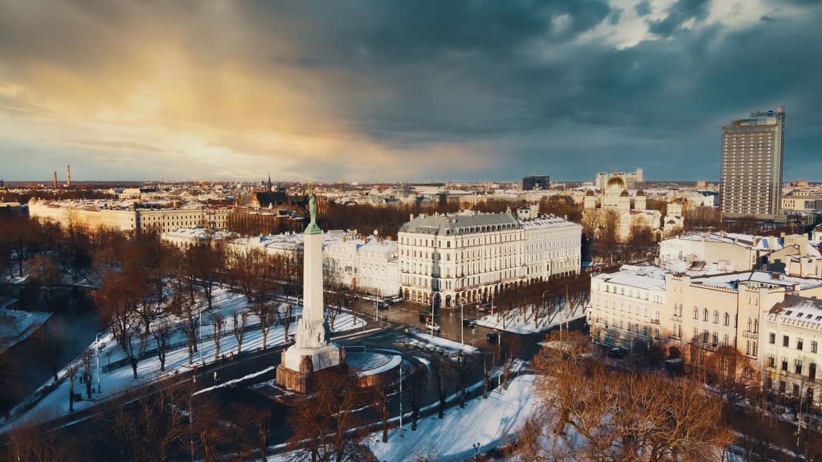Luftaufnahme des Freiheitsdenkmals in Riga, das markant auf einem Stadtplatz steht, umgeben von verschneiten Straßen und historischen Gebäuden im Sonnenuntergang.