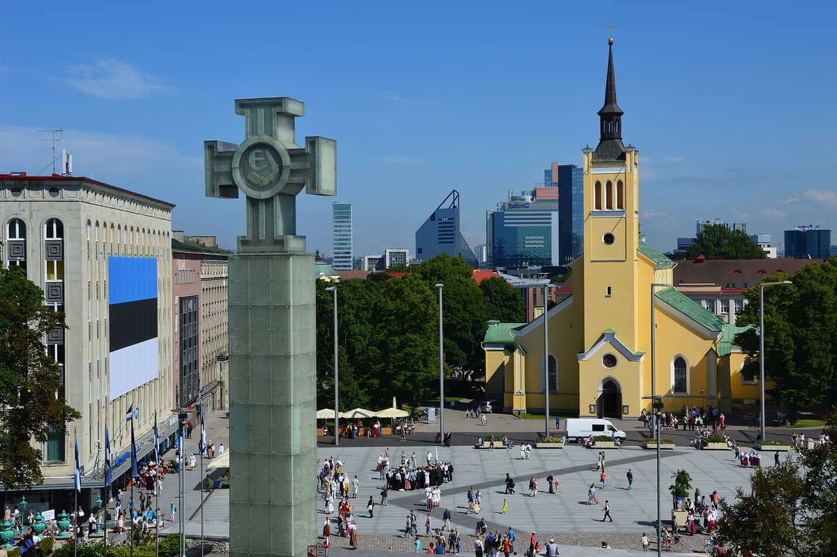 Der Freiheitsplatz in Tallinn mit der Siegessäule des Unabhängigkeitskrieges im Vordergrund, einer Kirche und dem Stadtpanorama im Hintergrund, Menschen auf dem Platz unter einem klaren blauen Himmel.