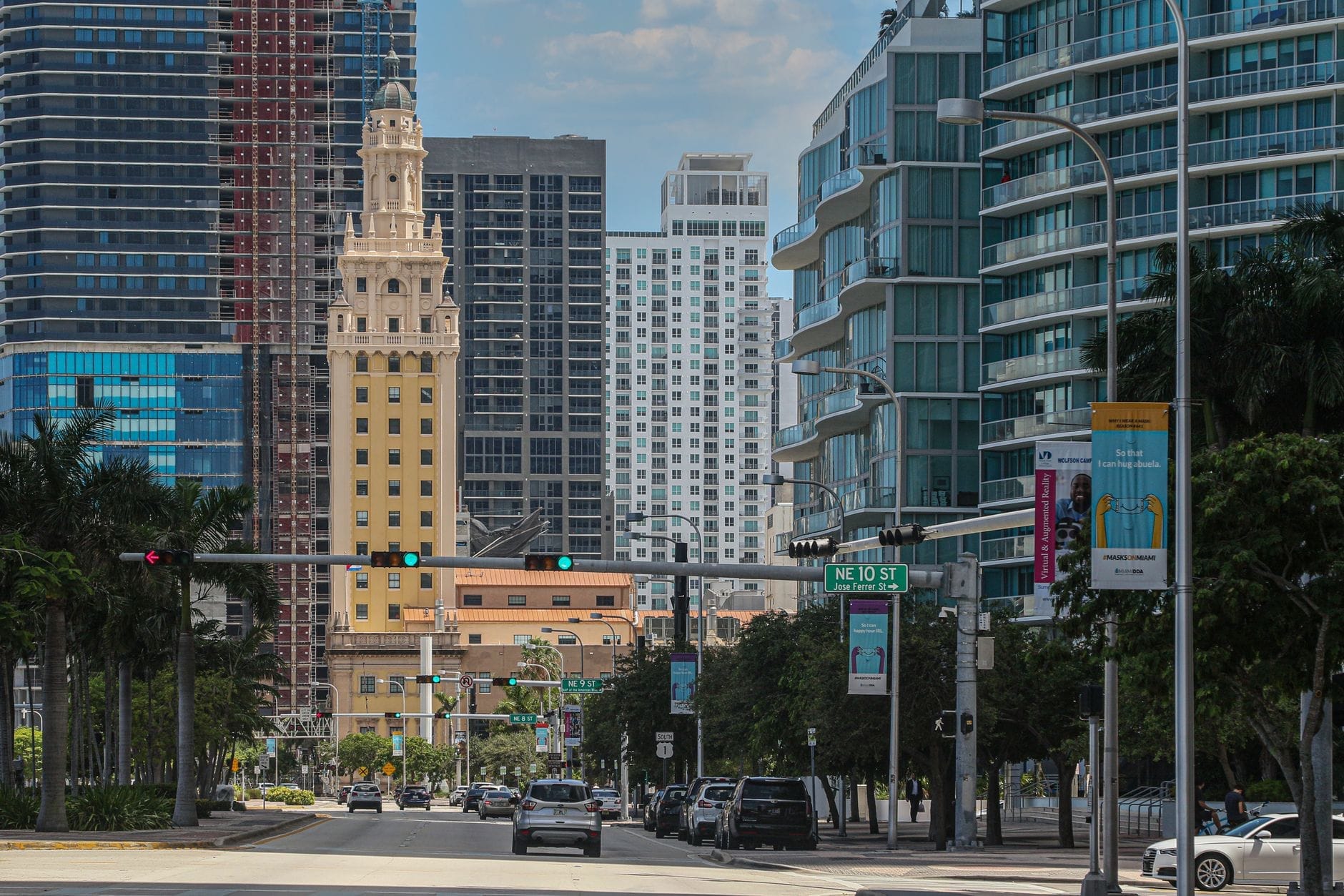 Il Freedom Tower campeggia al centro tra i grattacieli moderni di Miami, inquadrato lungo una vivace strada cittadina sotto un cielo luminoso, con traffico e palme che ne delimitano la scena.