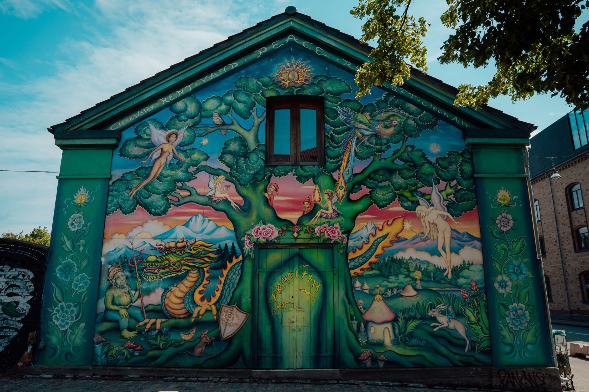Murales vivace sulla facciata di un edificio a Freetown Christiania, con una scena fantasy colorata che raffigura un albero, un drago, una fata ed elementi mistici, sotto un cielo azzurro.