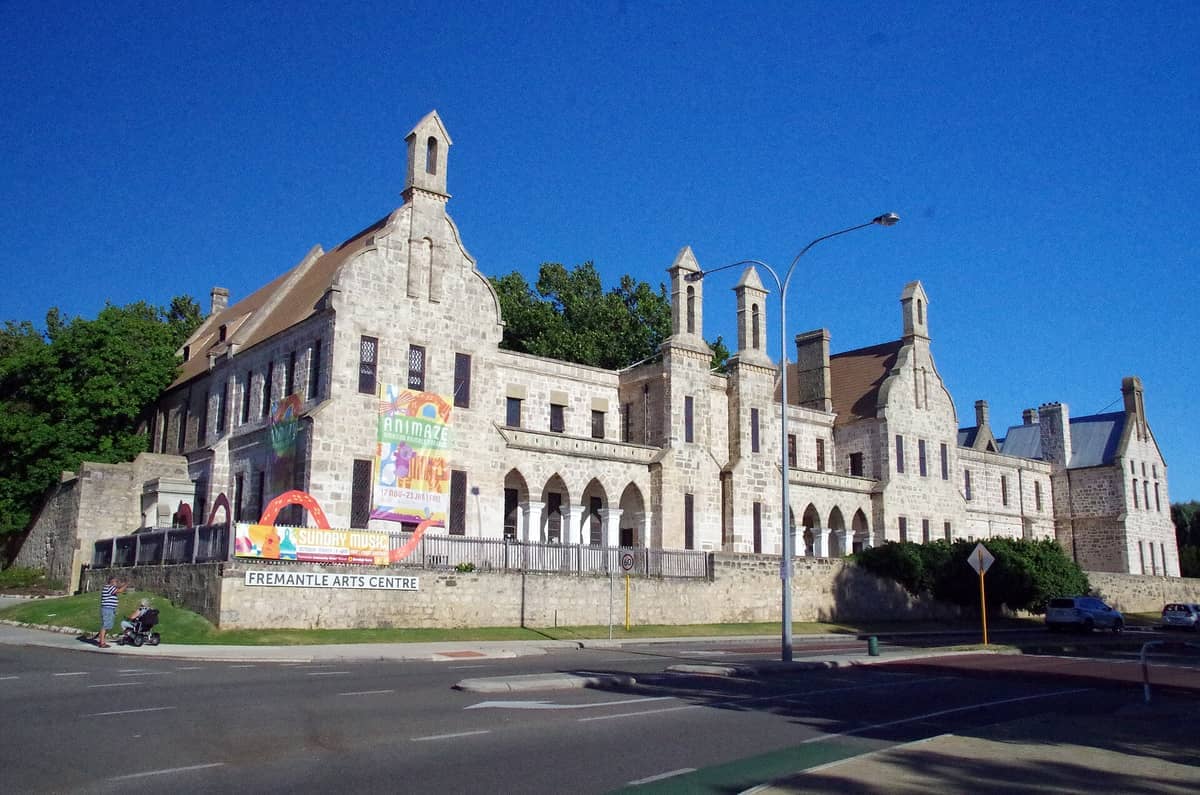 La imponente fachada gótica de piedra caliza del Walyalup Fremantle Arts Centre llena el encuadre, con vibrantes pancartas y un cielo azul despejado en lo alto.