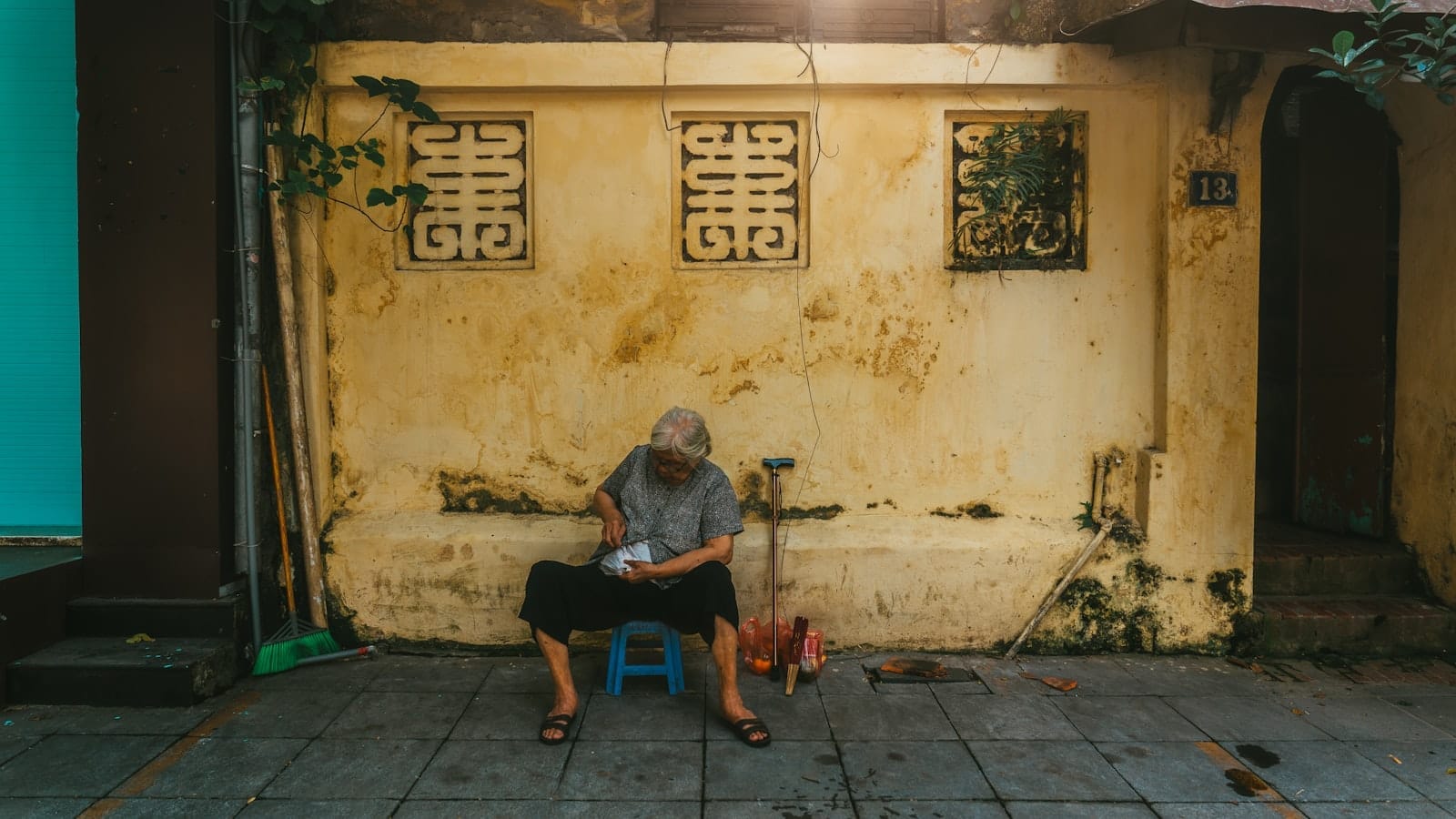 An elderly person sits on a blue stool by an ochre-colored wall with French architectural details, evoking the tranquil atmosphere of Hanoi's French Quarter.