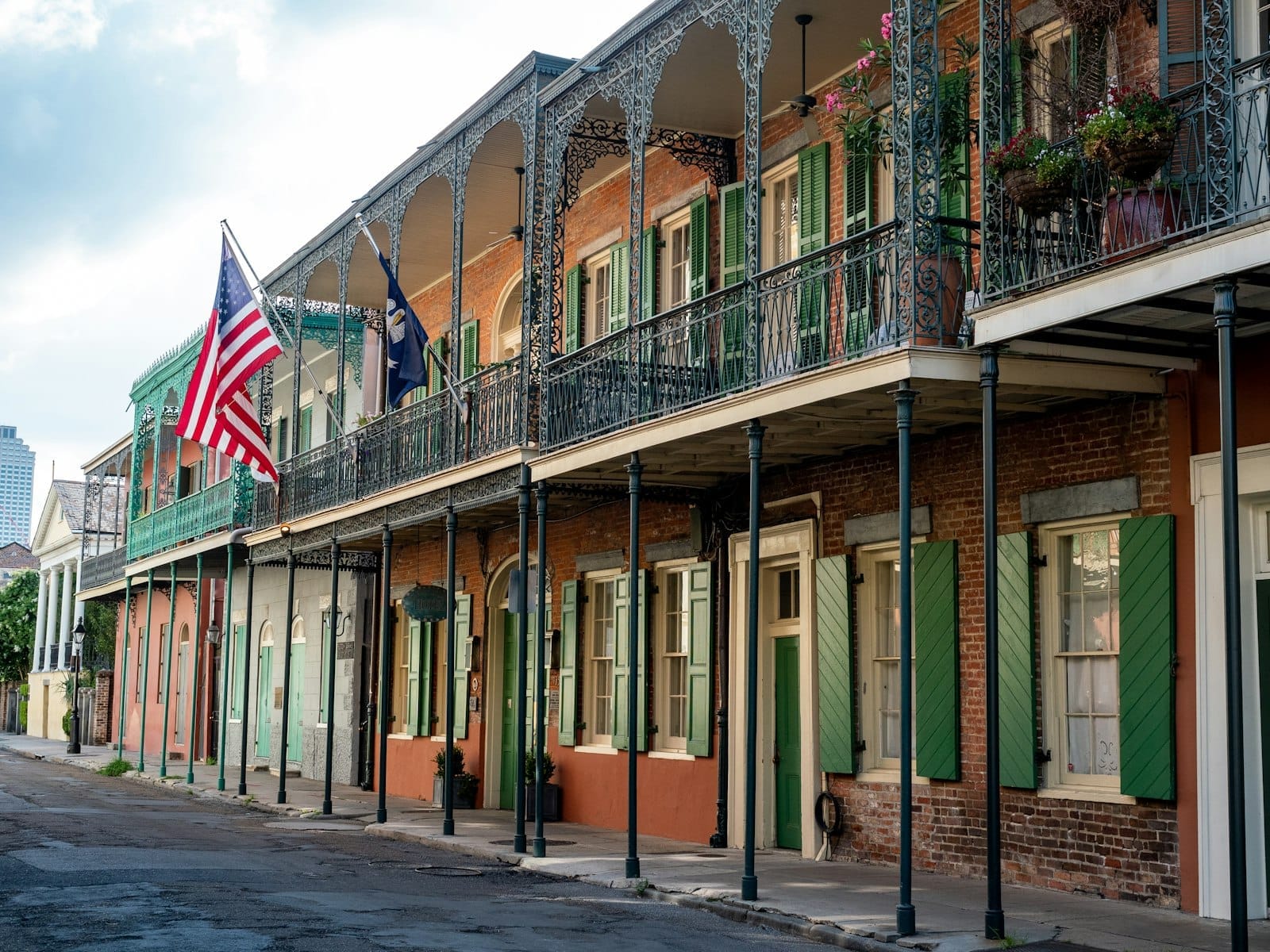 ภาพถนนใน French Quarter กับตึกรูปทรงสีสด ระเบียงเหล็กลาย หน้าต่างบานเกล็ดสีเขียว ธงชาติอเมริกัน และฟุตบาทสงบยามกลางวันอ่อน ๆ
