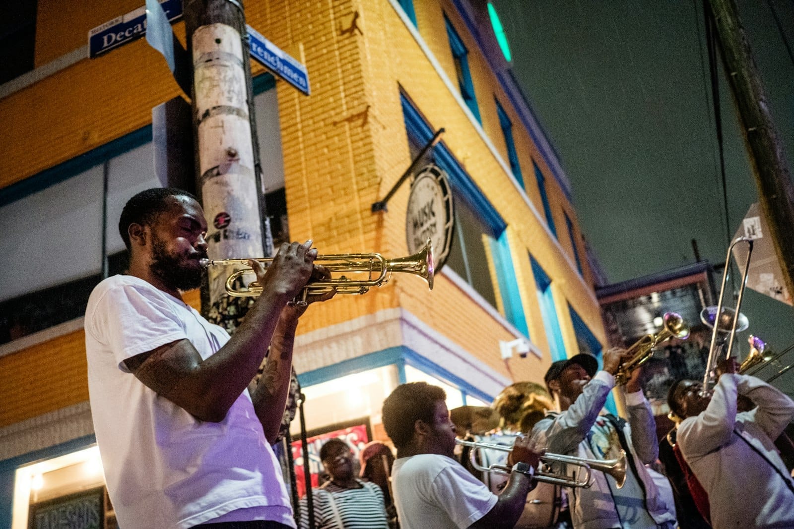 Musicisti suonano strumenti a fiato su Frenchmen Street di notte, con un edificio di mattoni gialli e cartelli stradali visibili sotto le luci della città.