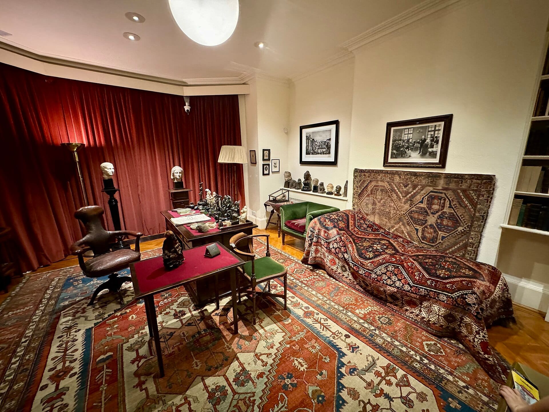 Sala delle consulenze del Freud Museum London con l'iconico lettino di Freud, tappeti ornati, scrivania antica e pareti ricche di opere d'arte in una luce calda.