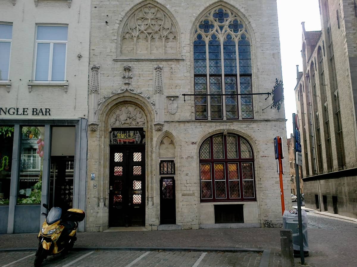 Facade of the Frietmuseum in Bruges showing its Gothic windows, ornate stonework, and entrance on Vlamingstraat, with a yellow scooter parked outside.