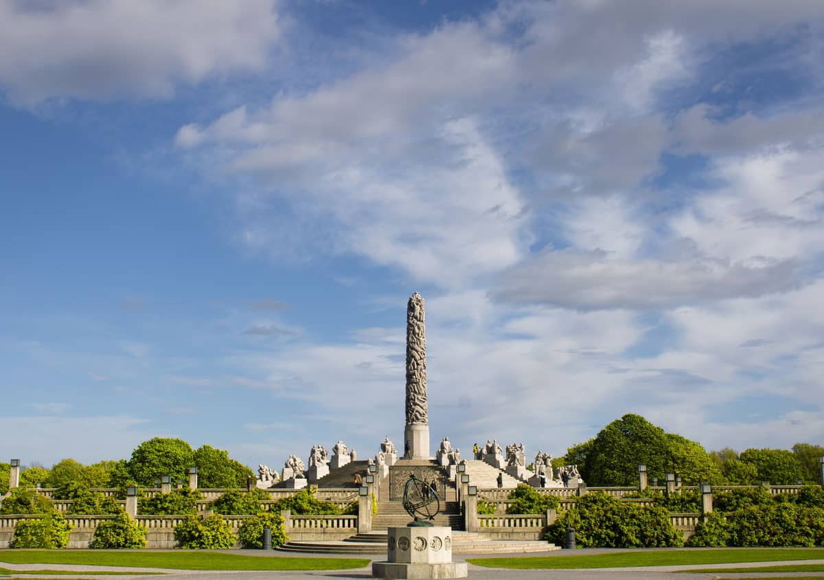 มุมกว้างของการจัดแสดงประติมากรรม Vigeland ในสวนฟร็อกเนอร์ เห็นเสา Monolith และรูปปั้นต่าง ๆ ล้อมรอบ ใต้ท้องฟ้าสีฟ้า ต้นไม้สีเขียวเป็นฉากหลัง
