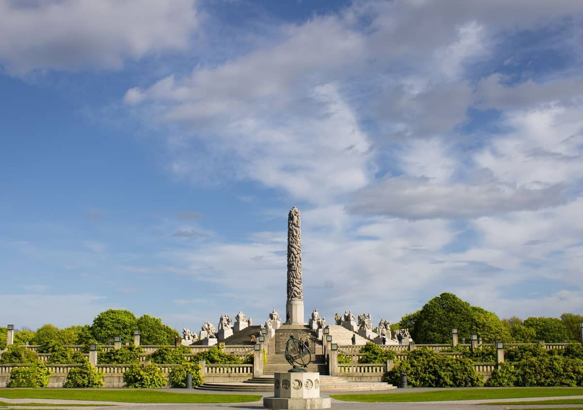 Monolith อันเป็นจุดเด่นกลางของ Vigeland Sculpture Park ล้อมรอบด้วยต้นไม้สีเขียว บันไดหิน และรูปปั้นใต้ท้องฟ้าสีฟ้ามีเมฆบางส่วนใน Frogner, Oslo