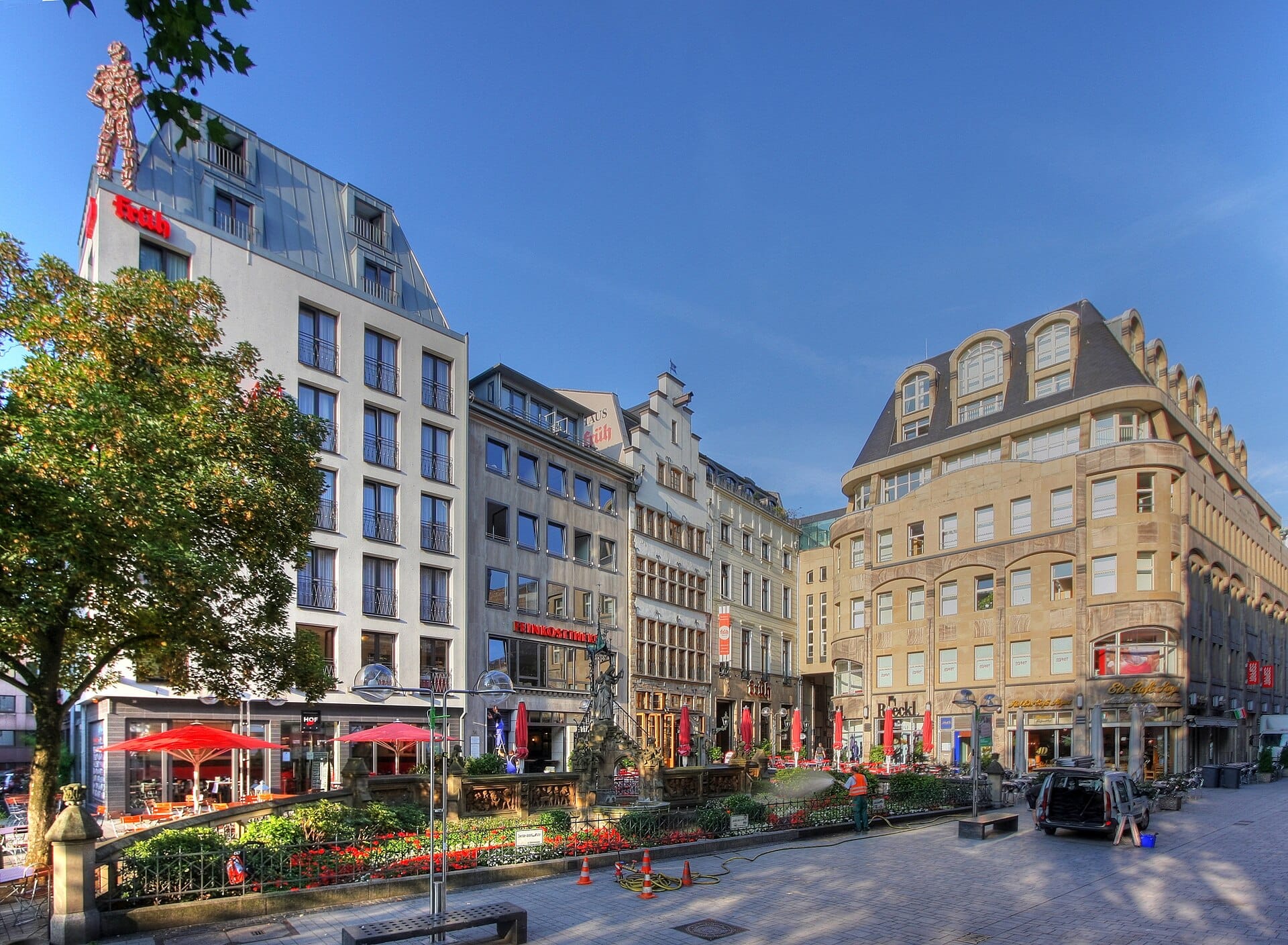 มุมกว้างของด้านหน้า Früh am Dom กับร่มสีแดง ที่นั่งด้านนอกข้างสวนเล็กๆ ใกล้มหาวิหารโคโลญจน์