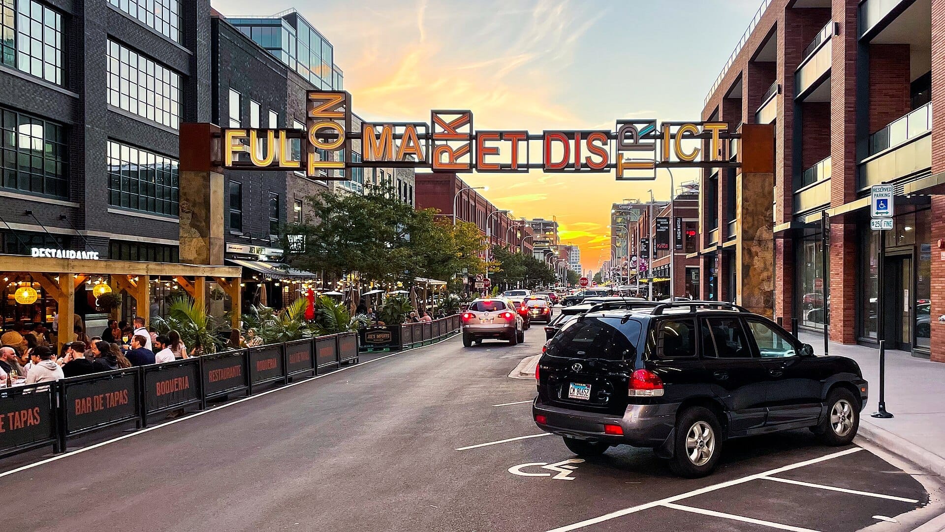 Insegna d'ingresso del Fulton Market District con una strada animata, edifici in mattoni, tavoli all'aperto, auto e un vivace cielo al tramonto a Chicago.