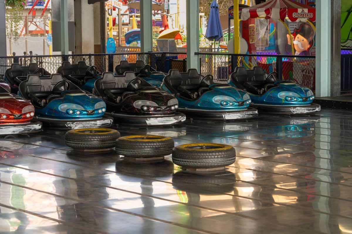 A row of colorful bumper cars lined up on a shiny indoor track with carnival rides and arcade games visible in the background.