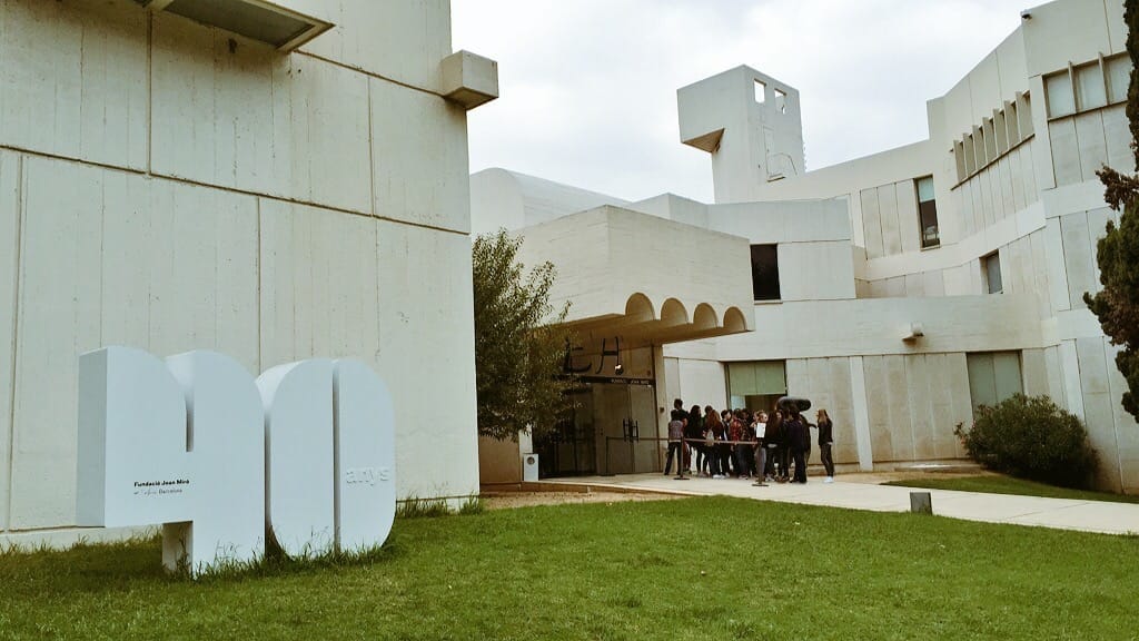 Exterior view of Fundació Joan Miró in Barcelona, showing modern white concrete architecture, grassy lawn, and a group of visitors near the entrance.