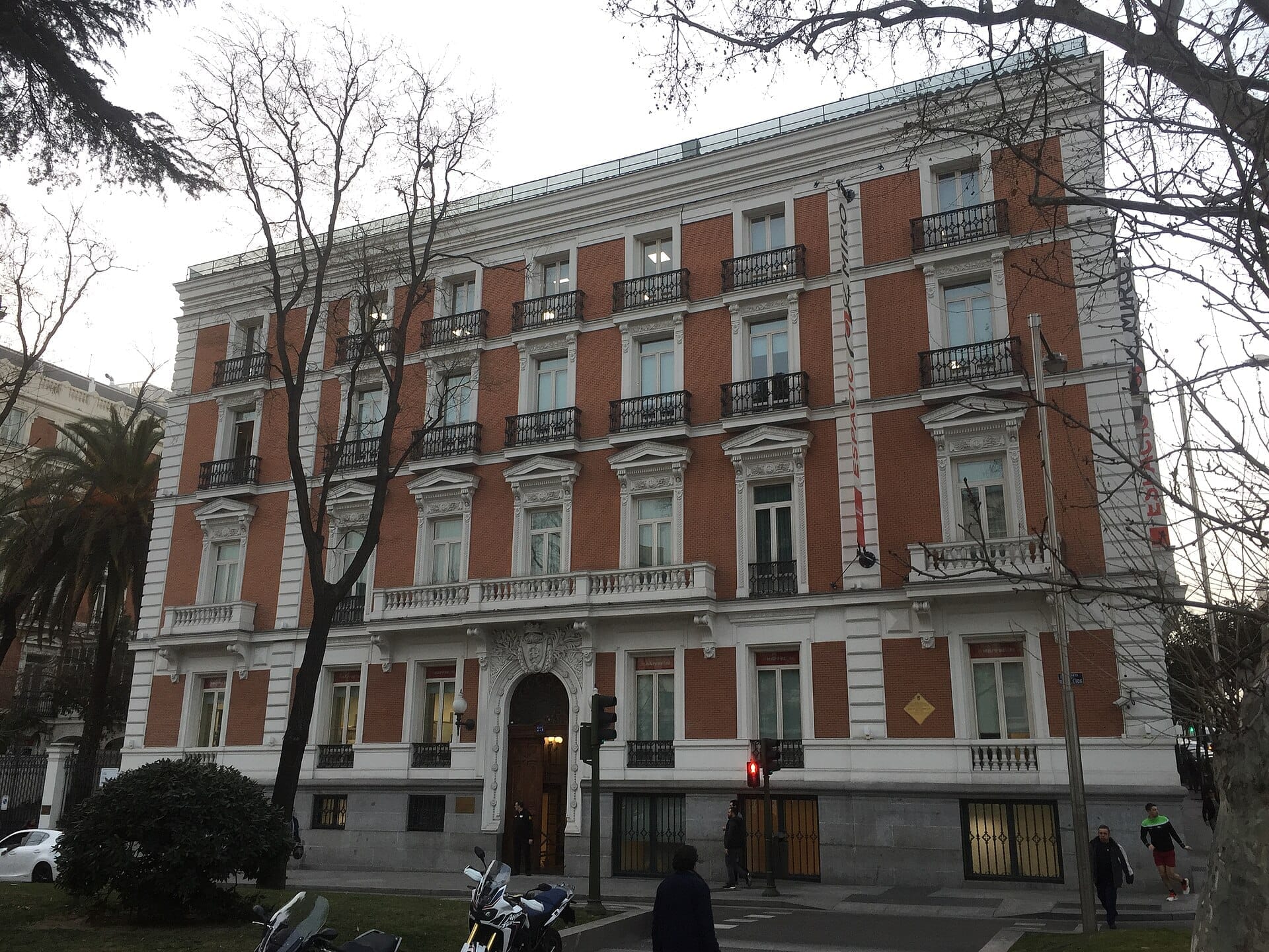 La Fundación MAPFRE Sala Recoletos presenta una maestosa facciata in mattoni rossi con raffinati dettagli bianchi, vista dall'altro lato di una tranquilla strada di Madrid.
