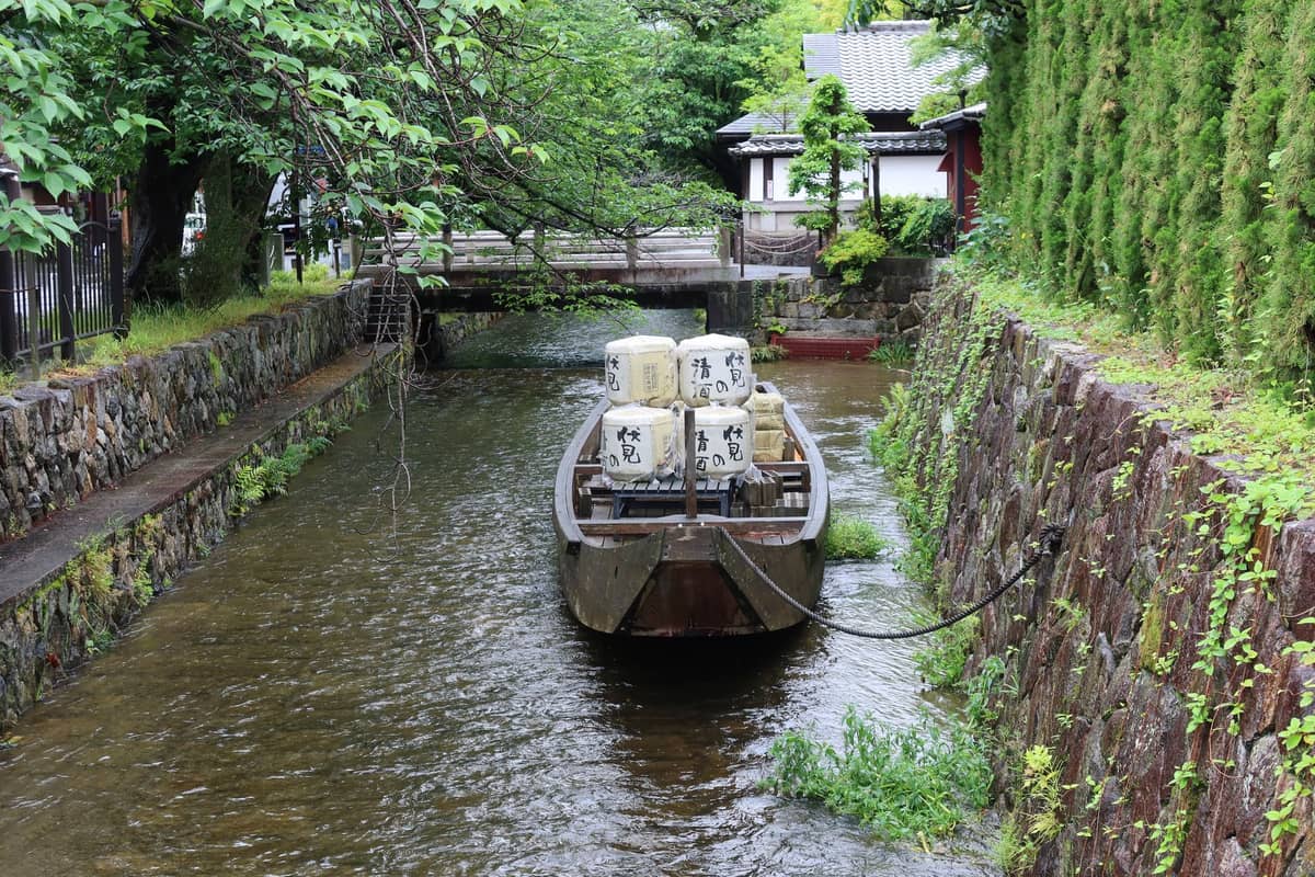 A traditional wooden boat carrying sake barrels floats on a quiet canal lined with stone walls and greenery in Kyoto’s Fushimi sake district.