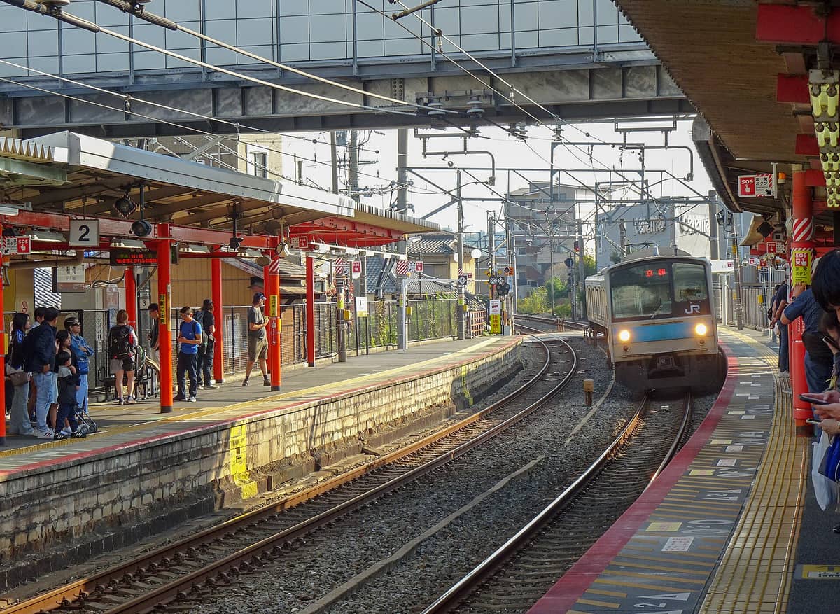 Wide view of JR Inari Station platform with orange beams, people waiting, and a local JR train approaching on the tracks in Kyoto.