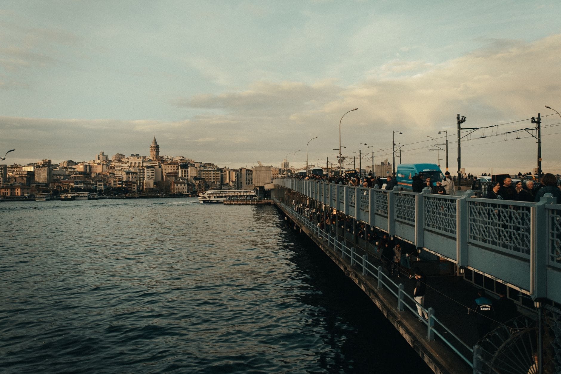 Il Ponte di Galata a Istanbul al crepuscolo, con persone che camminano, pescano e veicoli in transito, con lo skyline della città e la Torre di Galata sullo sfondo.