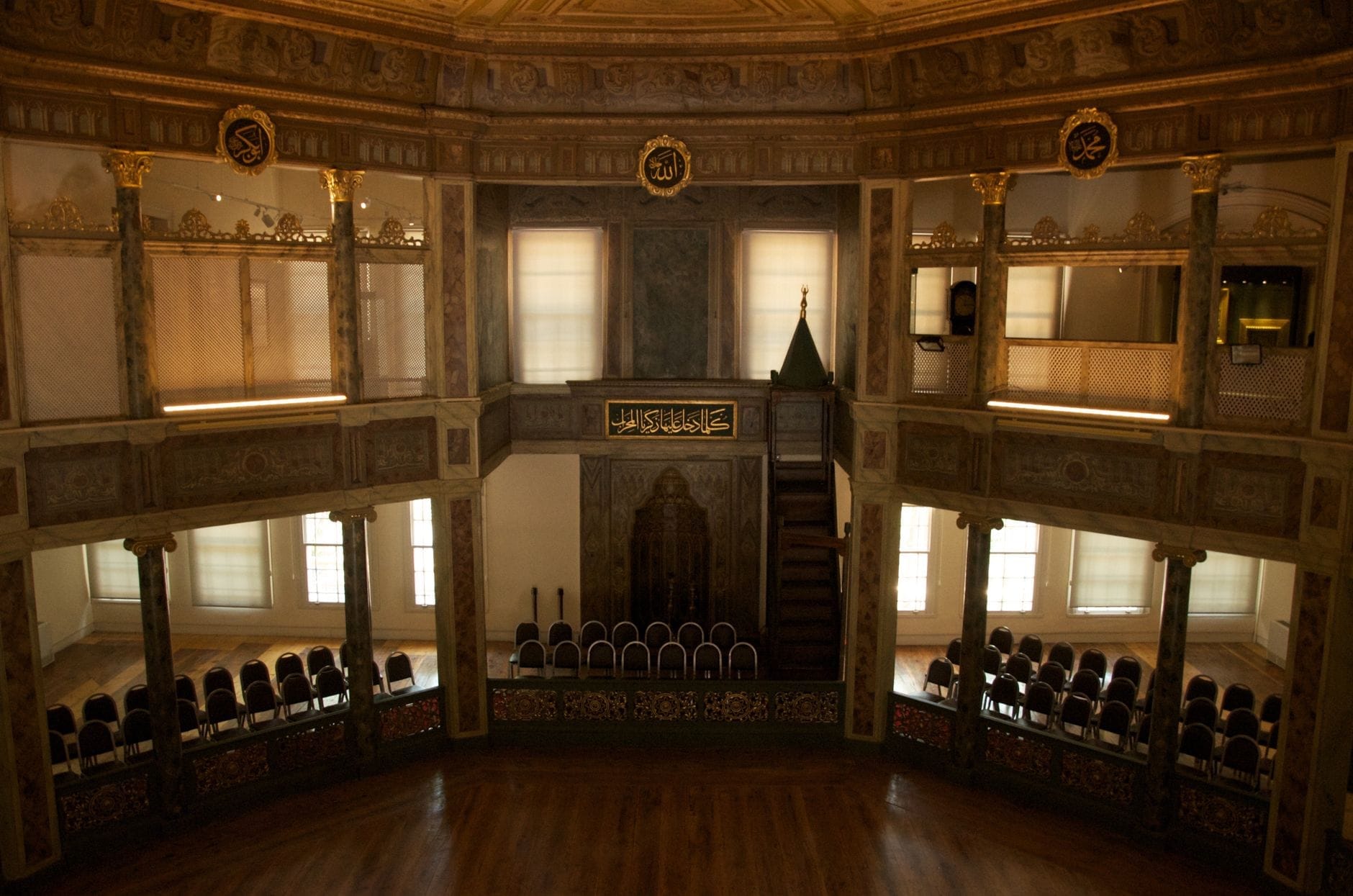 Interno del Museo Mevlevi di Galata con pavimenti in legno, balcone ornato, calligrafia decorativa e file di sedie vuote sotto la luce soffusa delle finestre.