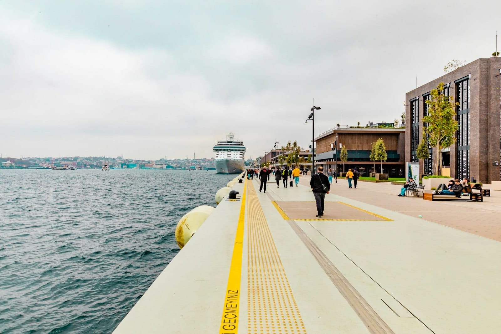 Passeggiata sul lungomare di Galataport Istanbul con persone che camminano, edifici moderni sulla destra e una nave da crociera ormeggiata lungo il Bosforo in una giornata nuvolosa.