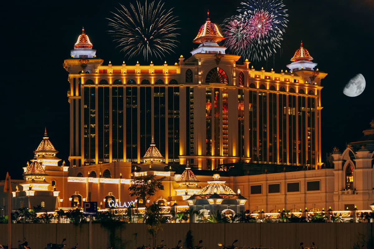 Nachtansicht der beleuchteten Hoteltürme des Galaxy Macau mit Feuerwerk am Himmel und dem über dem Resort sichtbaren Mond.