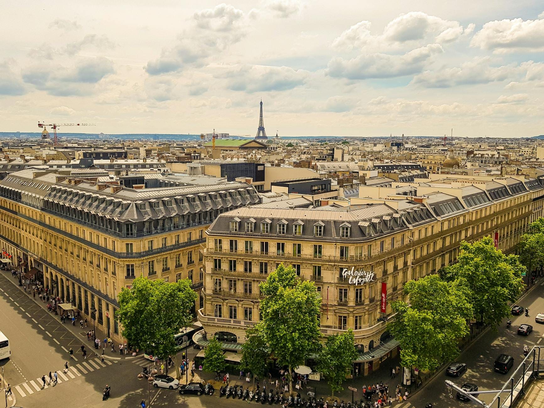 Vista aerea ampia dell'esterno delle Galeries Lafayette Haussmann con la classica architettura parigina, il viale alberato e la Torre Eiffel sullo sfondo sotto un cielo spettacolare.