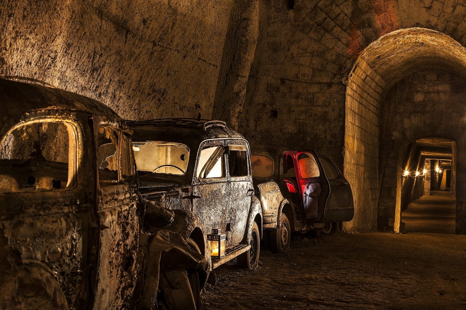 Auto arrugginite e abbandonate in fila nel tunnel in pietra scarsamente illuminato all'interno della Galleria Borbonica, con archi e pareti in tufo dalla texture visibile.