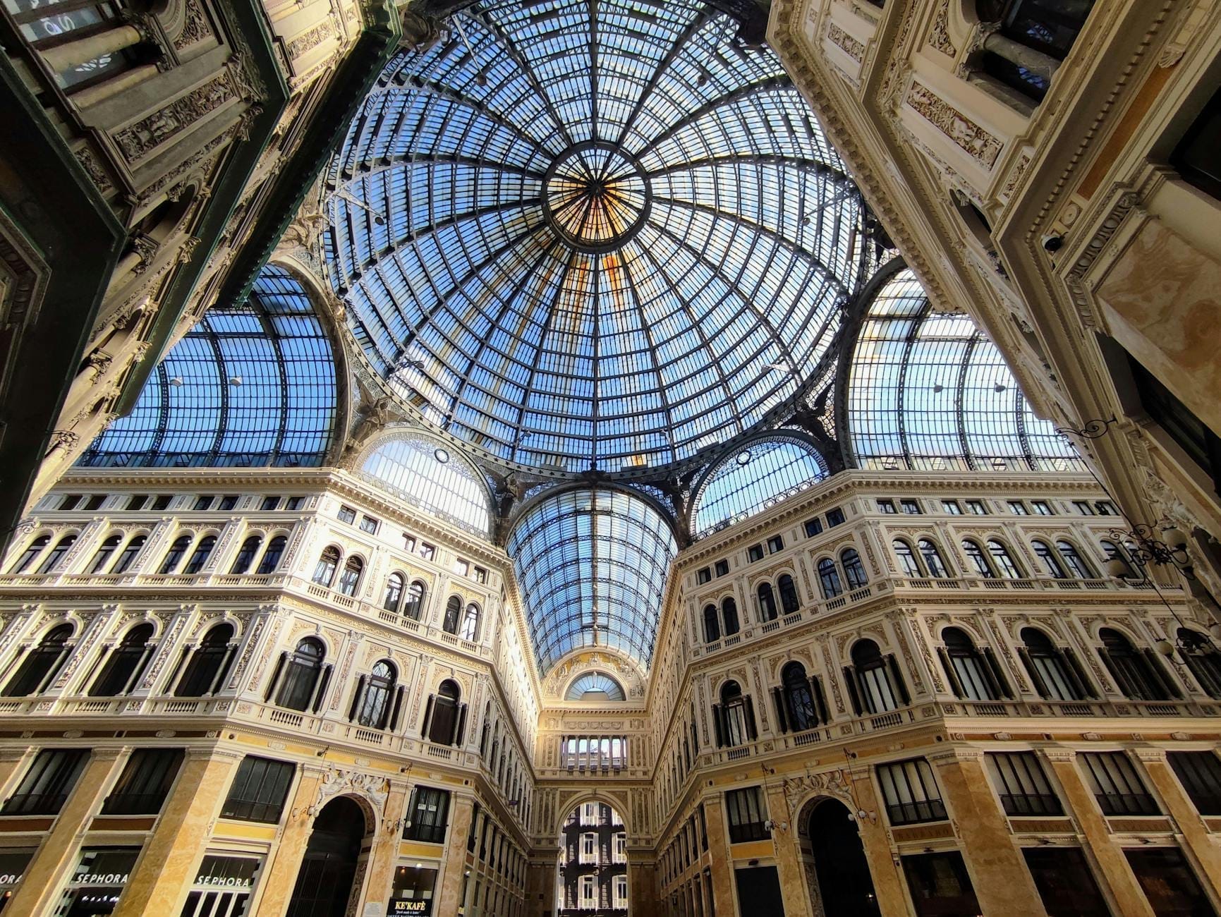 Vista grandangolare della maestosa cupola in vetro della Galleria Umberto I e della sua raffinata architettura ottocentesca, con la luce del sole che filtra attraverso le imponenti volte.