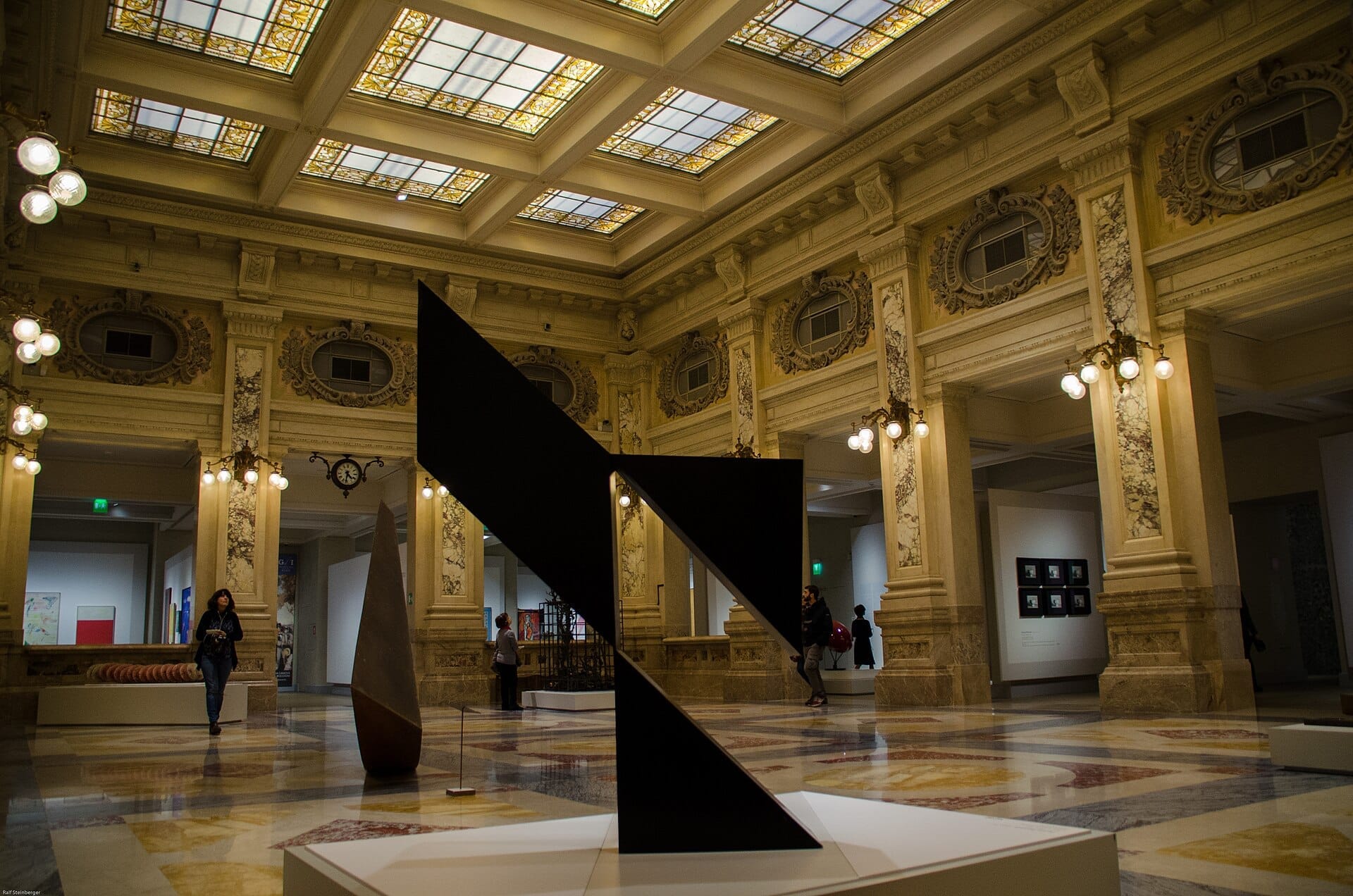 I visitatori esplorano il grande salone in marmo e le sculture moderne delle Gallerie d'Italia – Piazza Scala a Milano, sotto ornati soffitti di vetrate colorate.