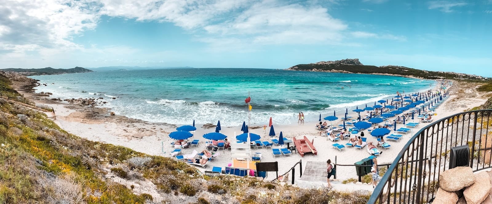 Panoramablick auf den Strand von Gallura auf Sardinien mit türkisfarbenem Wasser, weißem Sand, felsiger Küste und Reihen von blauen Sonnenschirmen und Liegestühlen.