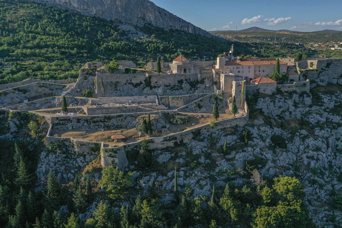 Luftaufnahme der Festung Klis auf einem felsigen Hügel, umgeben von Steinmauern und historischen Gebäuden, mit grünen Hügeln im Hintergrund.