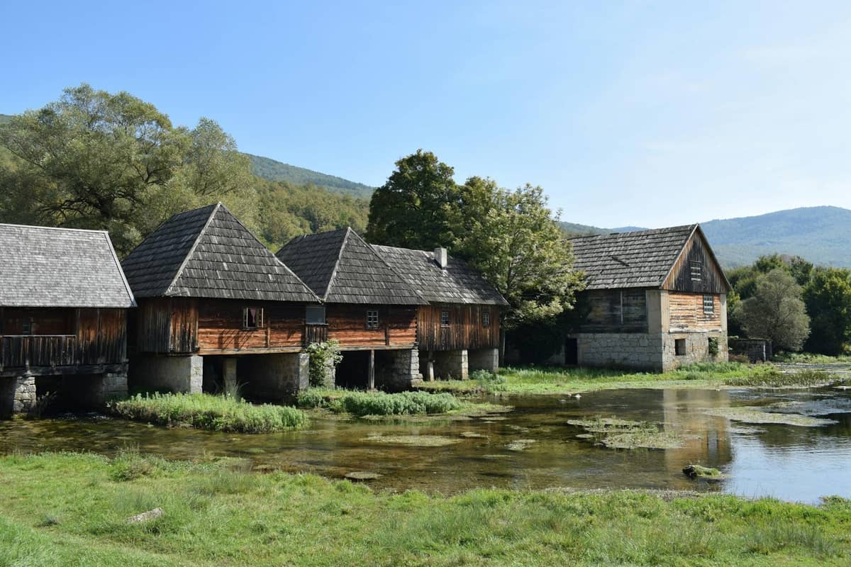 Reihe alter hölzerner Flussmühlen mit Steinsockeln entlang eines grasbewachsenen Flussufers unter klarem Himmel in der Nähe von Split, Kroatien.