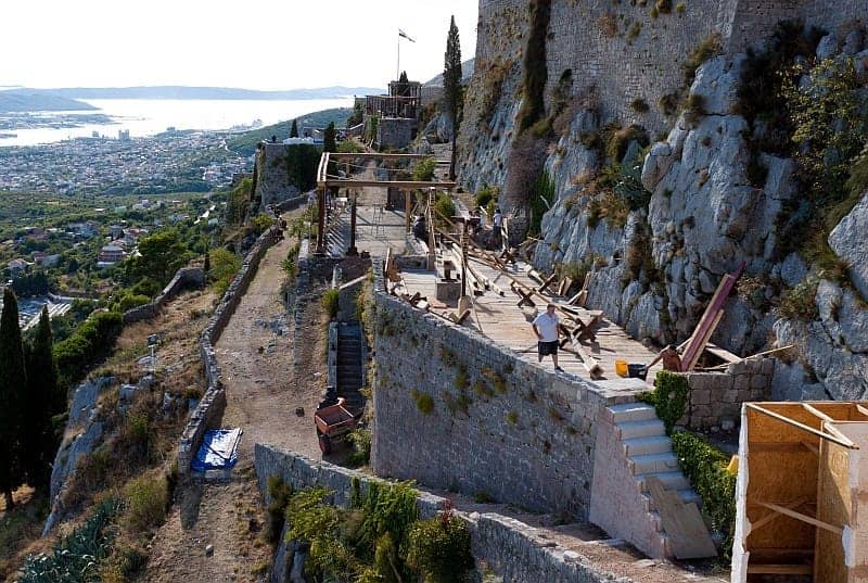 Malerischer Blick auf die Festung Klis in Kroatien mit Steinmauern, schroffen Klippen, Filmset-Konstruktionen und der in der Ferne sichtbaren Stadt Split.