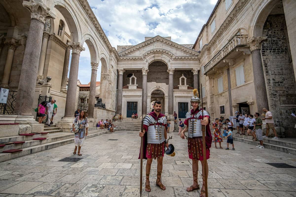 Weitwinkelansicht des Peristylplatzes in Split mit römischer Architektur und zwei kostümierten Wachen in Rüstungen, Touristen erkunden den historischen Kathedralenhof.
