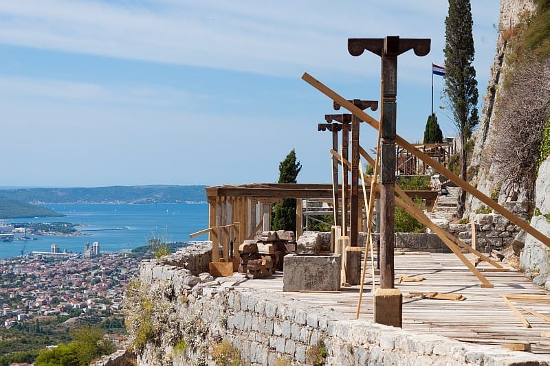 Estruturas de madeira de cenário de filmagem ao longo das muralhas da Fortaleza de Klis, com vista para a cidade de Split e a costa do Adriático sob um céu azul limpo.