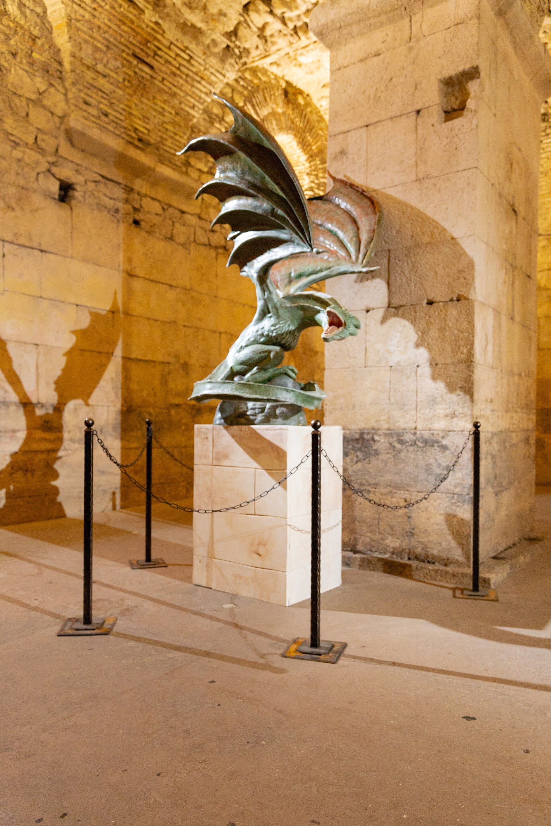 ประติมากรรมมังกรที่จัดแสดงบนแท่นภายใน Game of Thrones Museum Split ที่มีผนังหิน ล้อมรอบด้วยโซ่ตรวนและแสงไฟที่โดดเด่น