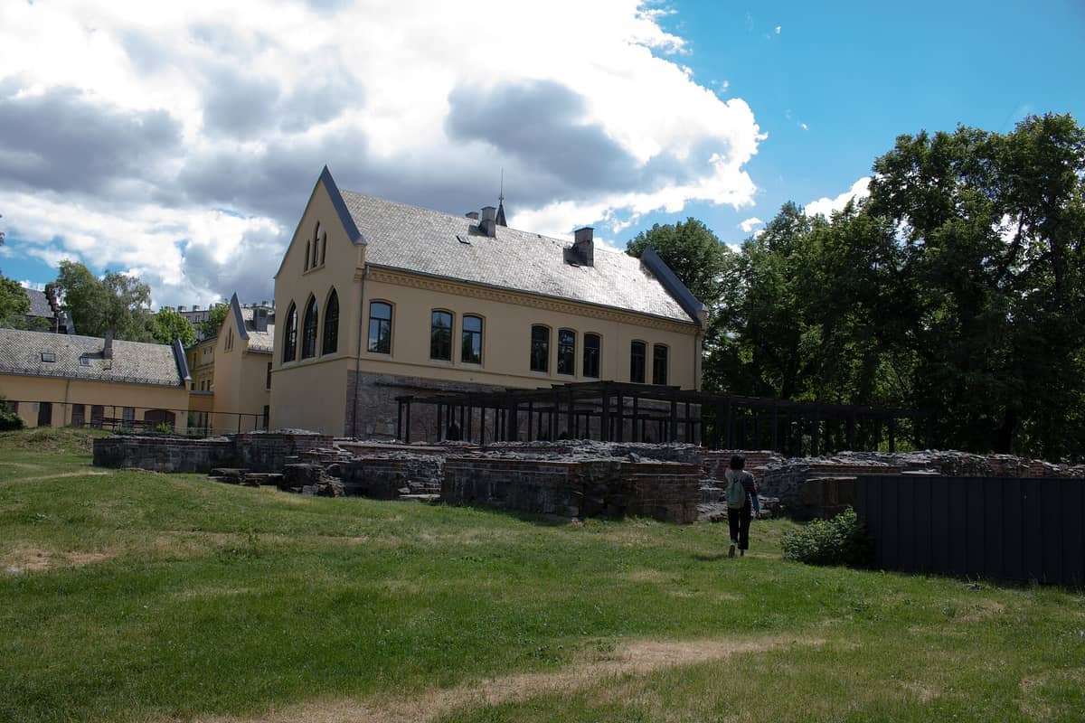 อาคารประวัติศาสตร์สีเหลืองและซากปรักหักพังหินยุคกลางใน Gamlebyen, Oslo ภายใต้ท้องฟ้าที่มีเมฆบางส่วนพร้อมต้นไม้สีเขียวล้อมรอบบริเวณนั้น
