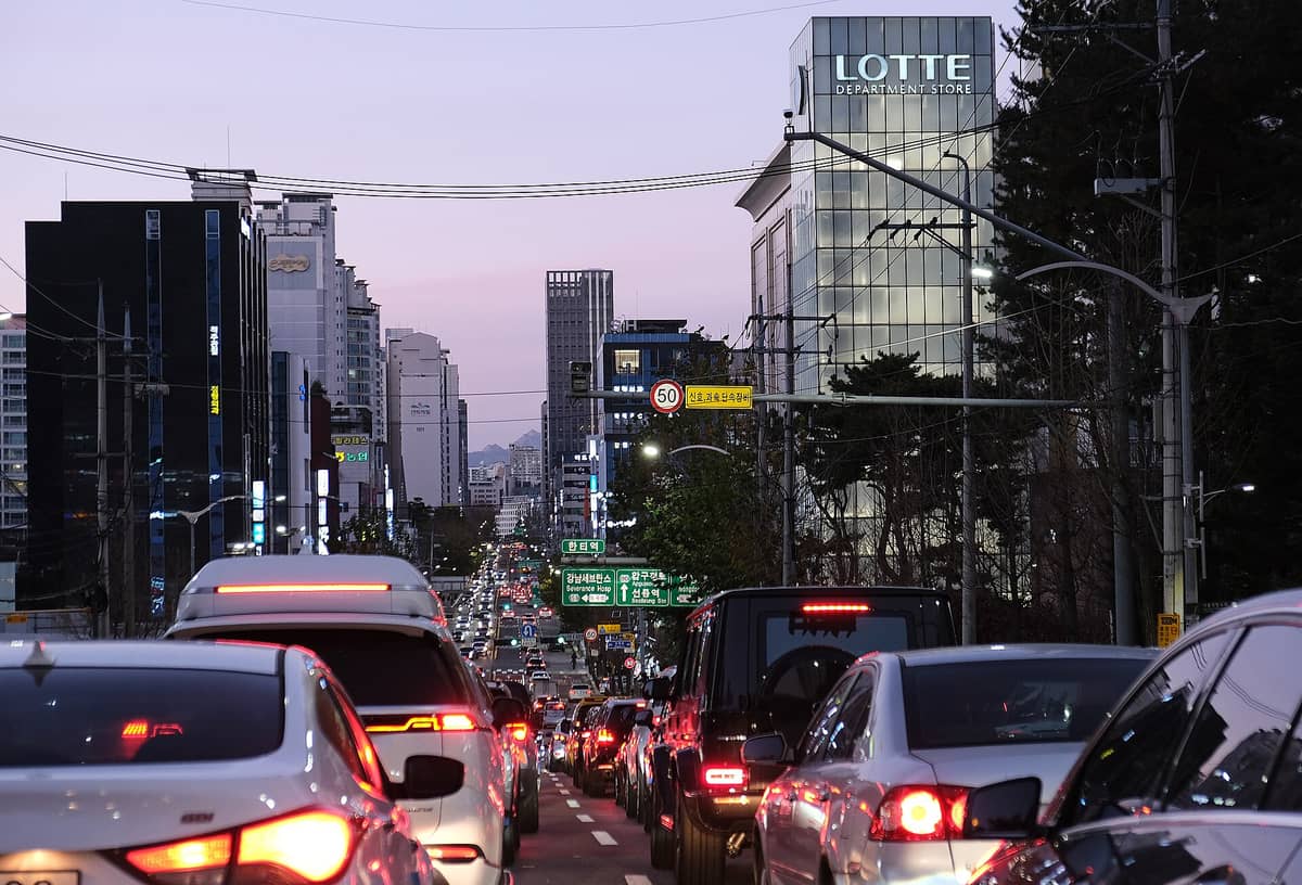 Cena de rua em Gangnam ao entardecer, com trânsito intenso, prédios de escritórios envidraçados, uma loja Lotte Department Store e placas de trânsito iluminadas no bairro Gangnam de Seul.