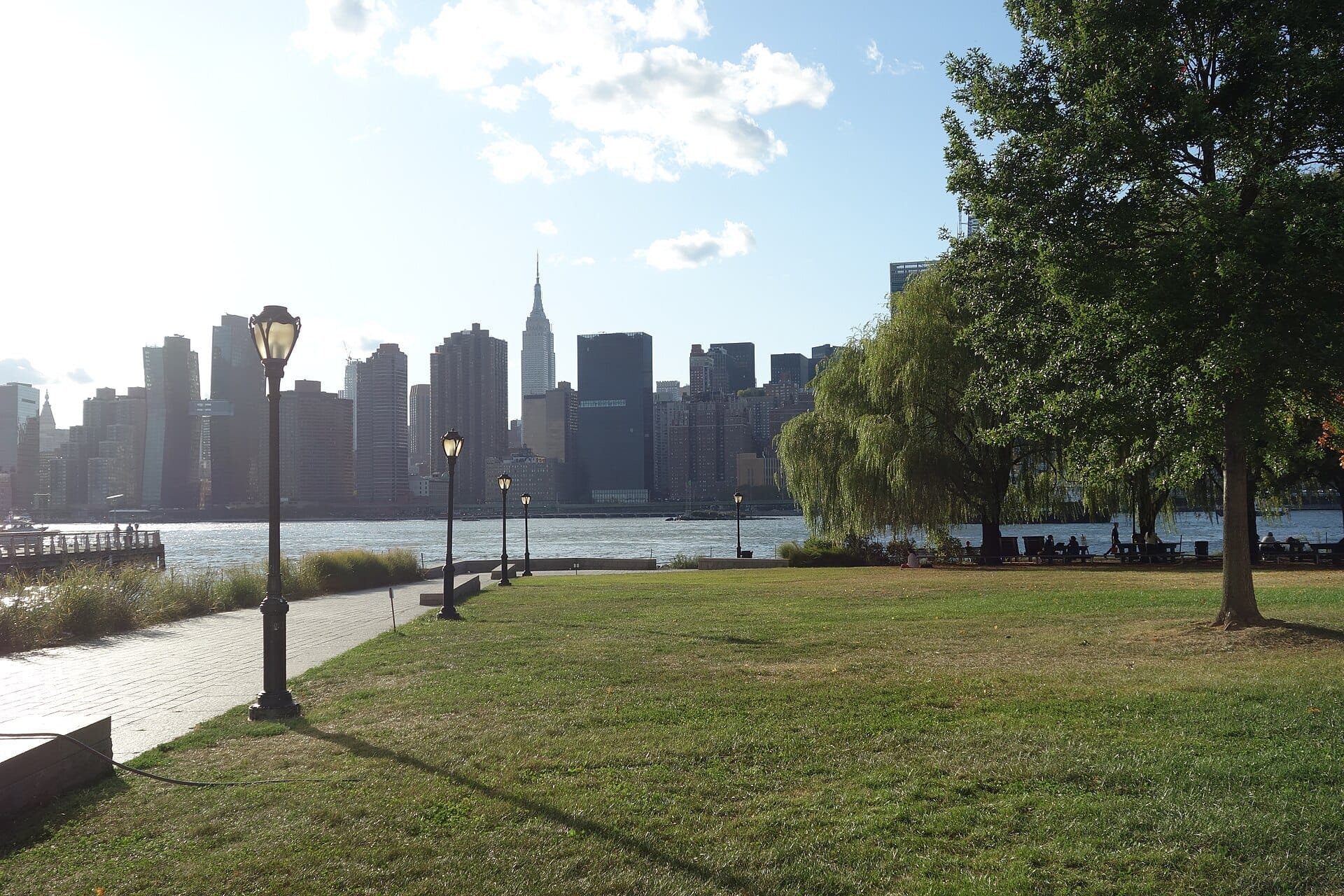 Prati verdi rigogliosi, ombra degli alberi e lampioni classici costeggiano il percorso del Gantry Plaza State Park con lo skyline di Midtown Manhattan che si erge sull'altra sponda dell'East River.